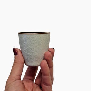 Op de afbeelding: Een kleine, gebroken witte keramische beker met een textuur en een donkerbruine rand. De beker wordt in een hand gehouden tegen een witte achtergrond. De beker is ongeveer 7.6 cm hoog.