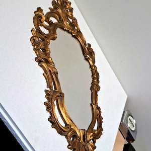 May include: Ornate, gold-colored oval mirror with a detailed, scrolling frame. The mirror has a reflective surface and is set against a white background. The frame features floral and leaf-like embellishments, creating a vintage aesthetic.