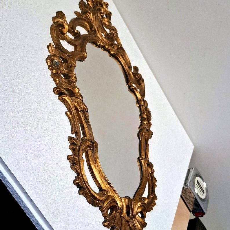 May include: Ornate, gold-colored oval mirror with a detailed, scrolling frame. The mirror has a reflective surface and is set against a white background. The frame features floral and leaf-like embellishments, creating a vintage aesthetic.