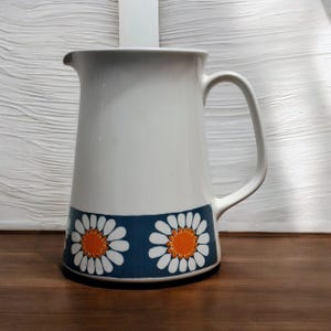 Può includere: Brocca in ceramica bianca con un manico grande e un beccuccio. La brocca presenta una fascia blu con margherite bianche e centri arancioni. La brocca è su una superficie di legno.