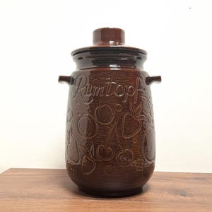 Op de afbeelding: Een bruine keramische Rumtopf-pot met een deksel en handvatten. De pot is versierd met in reliëf aangebrachte fruitontwerpen, waaronder aardbeien, kersen en perziken. Het woord "Rumtopf" staat in cursief geschreven.