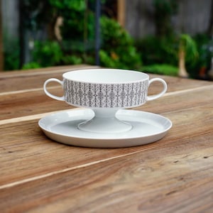 May include: White ceramic soup bowl with two handles, a pedestal base, and a matching saucer. The bowl has a black and grey geometric pattern around the rim. The set is designed for serving soup or other dishes.