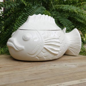 Könnte beinhalten: Eine weiße, fischförmige Servierschale aus Keramik mit Deckel. Die Schale hat detaillierte Schuppen, Flossen und einen Schwanz. Der Deckel ist wie die Rückenflosse des Fisches geformt. Die Schale steht auf einer Holzoberfläche.