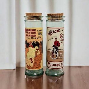 Könnte beinhalten: Zwei Vintage-Glasflaschen mit Korken. Eine zeigt ein Moulin Rouge-Postermotiv, die andere einen Radfahrer. Die Flaschen haben einen transparenten Glaskörper und dienen wahrscheinlich dekorativen Zwecken.