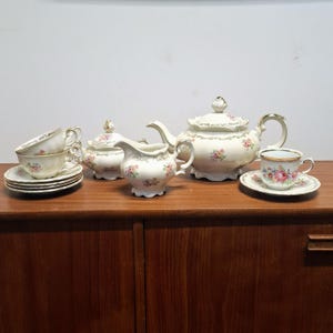 Può includere: Un servizio da tè vintage color crema con motivo floreale. Il set comprende una teiera, zuccheriera, lattiera, tazze e piattini. I bordi dei pezzi sono rifiniti con bordi dorati.