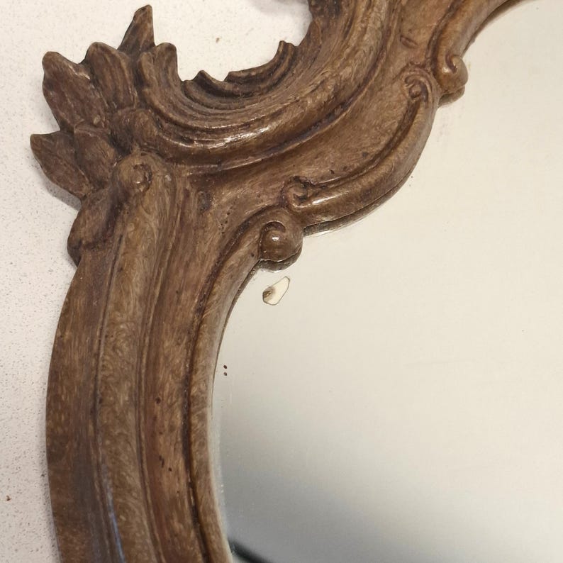 Puede incluir: Primer plano de un espejo decorativo con un marco de madera claro y ornamentado. El marco presenta tallas detalladas y un acabado brillante. La superficie del espejo refleja un fondo neutro.