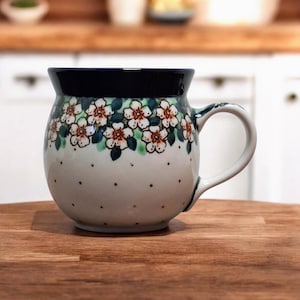 May include: A ceramic mug with a rounded body and a dark blue rim. The mug is decorated with a floral pattern of white and brown flowers with green leaves. The handle is white, and the mug sits on a wooden surface.
