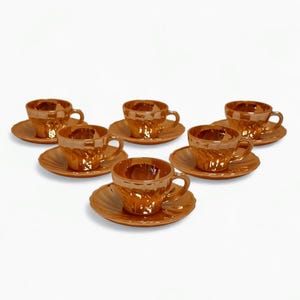 Op de afbeelding: Set van zes bijpassende oranje-bruine kopjes en schotels. Elk kopje heeft een oor en staat op een ronde schotel. De kopjes en schotels hebben een gestructureerd, geometrisch patroon. De set is op een wit oppervlak geplaatst.