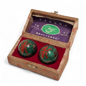 Op de afbeelding: Een set van twee groene marmeren oefenballen met rode Chinese karakters, gepresenteerd in een met stof gevoerde doos. De doos heeft een gouden sluiting en bevat een paars papier met Chinese tekst. Traditionele Chinese gezondheidsballen.