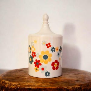 Può includere: Un vaso in ceramica color crema con coperchio, decorato con un motivo di fiori colorati in rosso, giallo, blu e bianco. Il vaso è cilindrico con una parte superiore arrotondata e poggia su una superficie di legno.