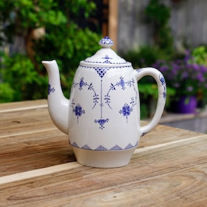 Könnte beinhalten: Weiße Keramik-Teekanne mit blauem Blumenmuster. Die Teekanne hat einen gebogenen Ausguss, einen großen Henkel und einen Deckel mit kleinem Knauf. Das blaue Design umfasst Blumen, Blätter und geometrische Muster.