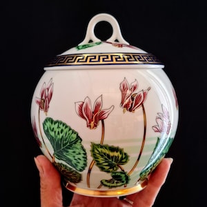 Può includere: Un barattolo rotondo in porcellana bianca con coperchio, decorato con un motivo dipinto a mano di fiori di ciclamino rosa e foglie verdi. Il coperchio ha un manico ad anello bianco e un bordo blu scuro con un motivo greco dorato.