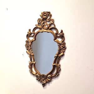 May include: Ornate gold-colored mirror with a decorative, floral-inspired frame. The mirror has a unique, curved shape and is mounted on a white wall. The frame features intricate carvings and embellishments, adding a touch of vintage elegance.