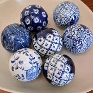 Może przedstawiać: Siedem niebiesko-białych ceramicznych kul dekoracyjnych z różnymi wzorami kwiatowymi i geometrycznymi. Kule ułożone są na białej, lekko zakrzywionej powierzchni. Wzory obejmują kwiaty, liście i skomplikowane linie.
