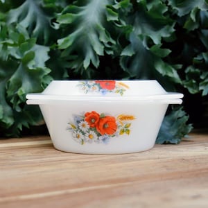 May include: A white, vintage casserole dish with a lid, decorated with a floral pattern of red poppies, daisies, and wheat. The dish is set against a backdrop of green foliage and a wooden surface.