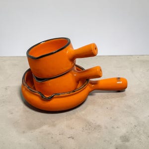 Puede incluir: Tres fuentes de cerámica naranja con asas. Los platos están apilados, con dos más pequeños encima de uno más grande. Cada plato tiene un borde negro. Las asas son cilíndricas y se extienden desde el lateral de cada plato.