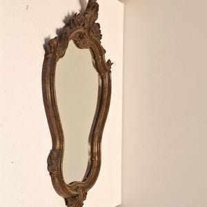 Puede incluir: Un espejo decorativo y ornamentado con un marco de madera marr&oacute;n. El espejo tiene una forma alargada y curvada con detalles tallados en la parte superior e inferior. El marco presenta un acabado envejecido, lo que le da un aspecto vintage.