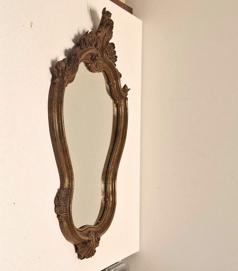Puede incluir: Un espejo decorativo y ornamentado con un marco de madera marr&oacute;n. El espejo tiene una forma alargada y curvada con detalles tallados en la parte superior e inferior. El marco presenta un acabado envejecido, lo que le da un aspecto vintage.