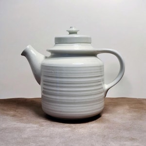 May include: A white ceramic teapot with a lid and a curved handle. The body of the teapot has horizontal ridged detailing. The teapot is sitting on a brown surface.