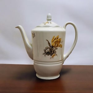 Può includere: Teiera in ceramica color crema con coperchio e manico curvo. La teiera è decorata con un motivo floreale con fiori gialli e marroni. Il beccuccio è lungo e curvo e il coperchio ha un piccolo pomello.