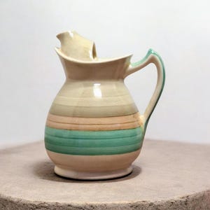 May include: A ceramic pitcher with a cream-colored spout and handle, and a body adorned with horizontal stripes in shades of beige, green, and peach. The handle is outlined in green. The pitcher sits on a neutral surface.