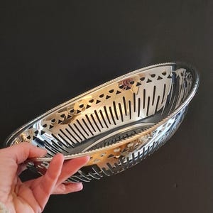 Può includere: Un cestino ovale argentato con un design traforato e bordo decorativo. Il cestino ha fessure verticali ed è tenuto da una mano. Lo sfondo è nero.