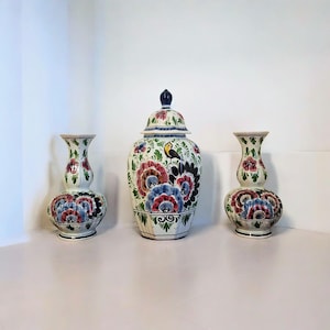 Könnte beinhalten: Drei Keramikvasen mit floralen Designs. Das Set enthält ein Gefäß mit Deckel und zwei kleinere Vasen. Die Vasen sind weiß mit roten, blauen und schwarzen Blumenmustern und grünen Blättern. Auf dem Gefäß ist ein gelber Vogel gemalt.