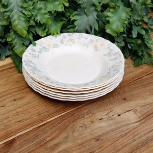 Könnte beinhalten: Ein Stapel von fünf weißen Keramik-Suppenschalen mit einem floralen Muster in Pastellfarben. Die Schalen haben einen gewellten Rand und sind auf einer Holzoberfläche mit grünem Laub im Hintergrund angeordnet.