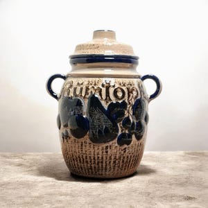 Op de afbeelding: Een vintage keramische pot met een deksel. Het heeft een getextureerde bruine basis en een beige bovenkant. De pot heeft donkerblauwe accenten, waaronder de handvatten en bloemmotieven. Het woord "Rümtop" staat op de pot.