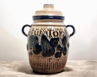 Peut inclure: Un pot en céramique vintage avec un couvercle. Il présente une base marron texturée et un dessus beige. Le pot a des accents bleu foncé, y compris les poignées et les motifs floraux. Le mot "Rümtop" est écrit sur le pot.