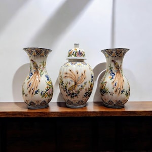 Può includere: Tre vasi in ceramica con un motivo a pavone. I vasi sono bianchi con un motivo dettagliato di pavone, fiori e fogliame nei toni del marrone, blu e giallo. I vasi sono su una superficie di legno scuro.