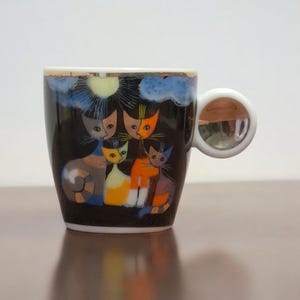 以下が含まれることがあります： 白いつまみ付きの小さな黒いセラミックコーヒーカップ。カップには、太陽と雲のデザインの下に、5匹の様式化された猫のカラフルなイラストが描かれています。猫はオレンジ、グレー、黄色、青の色合いです。