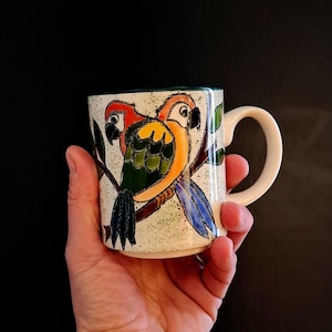 Könnte beinhalten: Keramiktasse mit einem bunten Papageien-Design. Die Papageien mit roten, gelben, grünen und blauen Federn sitzen auf einem Ast. Die Tasse hat einen cremefarbenen, gesprenkelten Hintergrund und einen gebogenen Henkel.