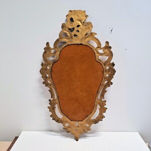 May include: Ornate gold-colored wall mirror with a decorative, scrolling frame. The mirror has a brown backing and a detailed, vintage design. The frame features intricate carvings and a distressed finish, adding to its antique aesthetic.