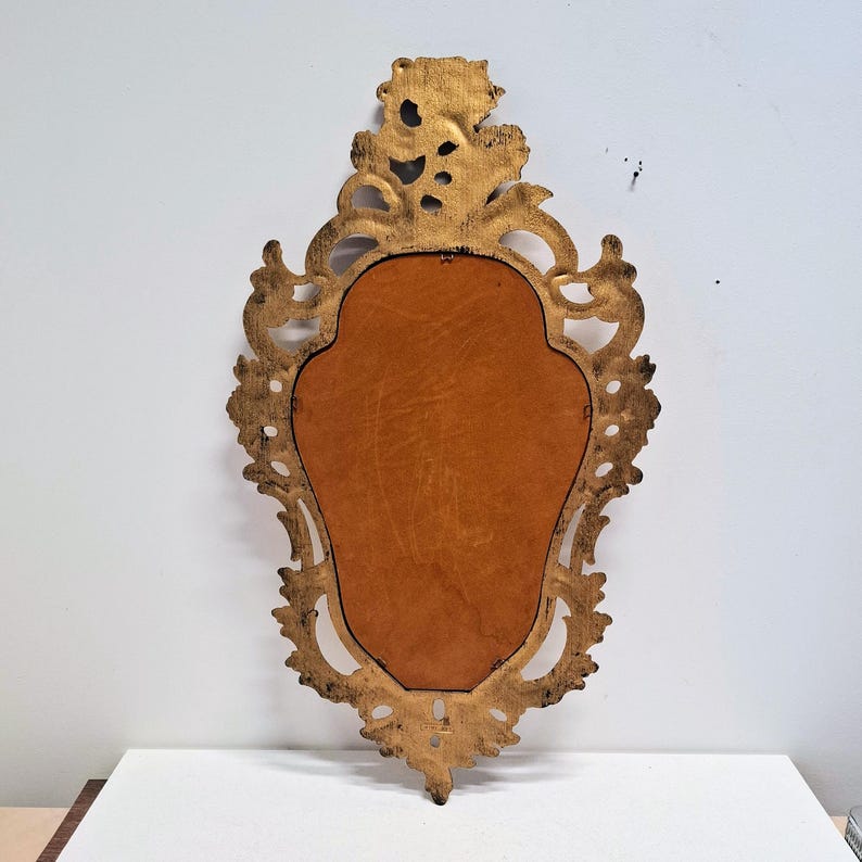 May include: Ornate gold-colored wall mirror with a decorative, scrolling frame. The mirror has a brown backing and a detailed, vintage design. The frame features intricate carvings and a distressed finish, adding to its antique aesthetic.