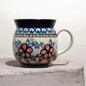 Può includere: Una tazza in ceramica con corpo arrotondato e manico. La tazza presenta un motivo floreale con fiori arancioni e blu su sfondo bianco. Il bordo è blu scuro e il manico è bianco.