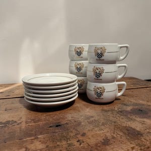Op de afbeelding: Een stapel witte keramische schotels met een gouden rand naast een stapel bijpassende koffiekopjes. De kopjes en schotels hebben een goud en zwart wapenontwerp en het woord "Hamboule". De items staan op een houten oppervlak.