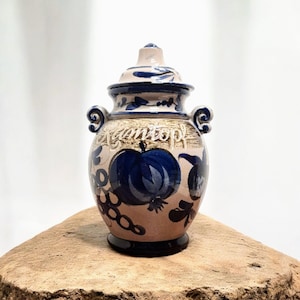 Op de afbeelding: Een keramische vaas met deksel, met een beige basis en blauwe accenten. De vaas is versierd met een blauw fruitontwerp en het woord "Camtop" in cursief. Hij heeft twee handvatten en staat op een houten oppervlak.