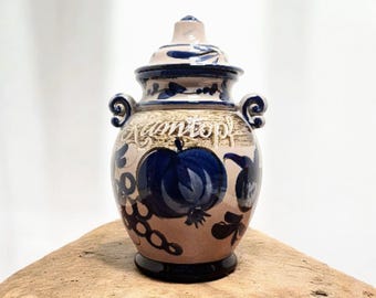 Peut inclure: Un vase en céramique avec couvercle, base beige et détails bleus. Le vase est orné d'un motif de fruit bleu et du mot "Camtop" en cursive. Il possède deux anses et repose sur une surface en bois.