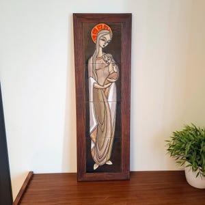 May include: A framed ceramic tile artwork depicting a religious figure holding a child. The artwork features a woman with a halo, dressed in flowing robes with a colour palette of beige, brown, and white. The frame is a dark wood.