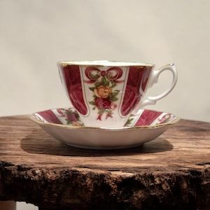 Peut inclure: Une tasse et une soucoupe vintage. La tasse et la soucoupe sont blanches avec un motif bordeaux et floral. La tasse a un bord et une anse dorés. La soucoupe a un motif bordeaux et floral assorti. L'ensemble est sur une surface en bois.