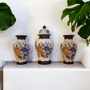 Op de afbeelding: Drie decoratieve keramische vazen met bloemmotieven. De vazen zijn beige met zwarte randen en bodems. Het bloemmotief bevat oranje, blauwe en groene bloemen met bruine bladeren. Eén vaas heeft een deksel.