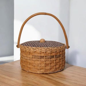 以下が含まれることがあります： 木製の持ち手と花柄の布の蓋が付いた、丸い柳のバスケット。バスケットは薄茶色で、蓋には濃い花柄模様があります。持ち手はアーチ型で木製です。