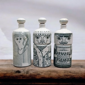 May include: Three white ceramic bottles with black illustrations. The bottles feature different face designs and are topped with white stoppers. One bottle has a woman's face, another a king's face, and the third has a label that says "SCOTCH WHISKY".