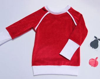 Op de afbeelding: Een rood velours shirt met lange mouwen en witte afwerking op de kraag, mouwen en zoom. Het shirt heeft een raglan mouwontwerp. Een zwarte peer en rode appel staan naast het shirt.