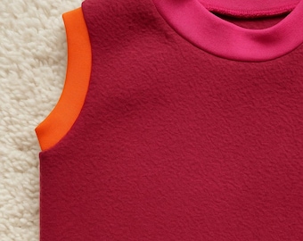 Op de afbeelding: Een mouwloze top in een dieprode kleur, met een contrasterende oranje afwerking langs de armsgaten en een felroze halslijn. De stof heeft een getextureerde uitstraling, wat wijst op een zacht, comfortabel materiaal. Het kledingstuk is ontworpen met een hoge halslijn.
