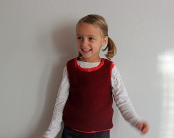 Op de afbeelding: Een jong meisje met blond haar draagt een rode, mouwloze trui over een wit shirt met kleine zwarte stippen. Ze glimlacht en kijkt naar rechts.