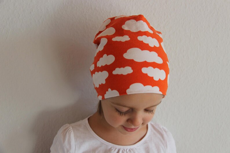 Puede incluir: Un ni&ntilde;o o ni&ntilde;a lleva una boina de color naranja brillante con un patr&oacute;n de nubes blancas. La boina est&aacute; hecha de un tejido suave y el&aacute;stico.
