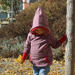Puede incluir: Una persona joven lleva una sudadera con capucha morada con ribetes rojos. La capucha está puesta sobre la cabeza. Está de pie en un patio con hojas caídas.