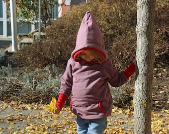Op de afbeelding: Een jong persoon draagt een paarse hoodie met rode bies. De capuchon is over het hoofd getrokken. Ze staan in een tuin met gevallen bladeren.